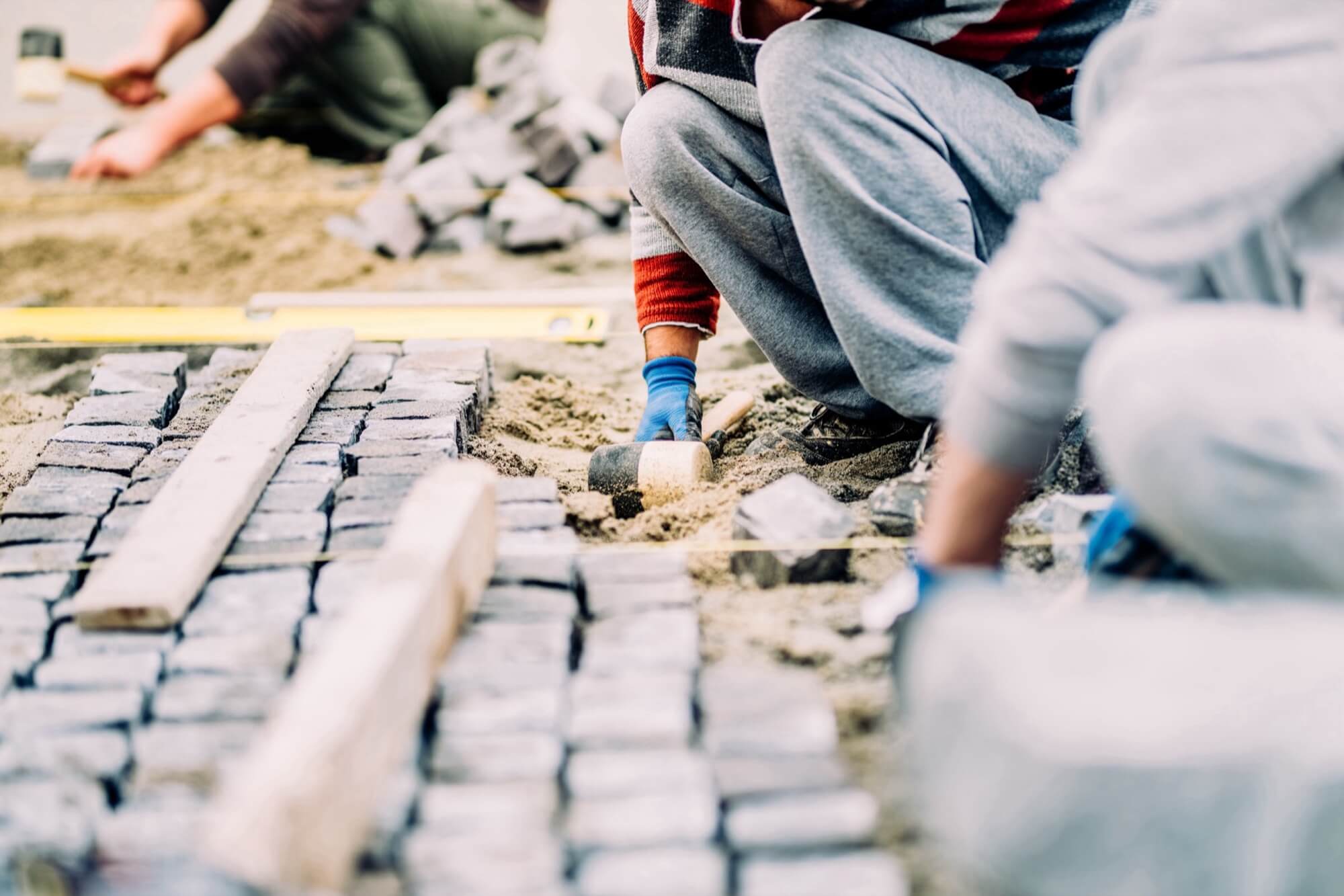 Workers in work clothes and gloves lay paving stones on a sand bed, with tools like a rubber mallet and spirit level visible.